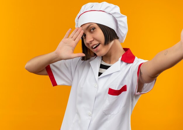 Photo gratuite joyeuse jeune femme cuisinier en uniforme de chef étendant la main et faisant des gestes salut isolé sur orange