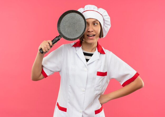 Joyeuse jeune femme cuisinier en uniforme de chef tenant et à la poêle à frire isolé sur rose