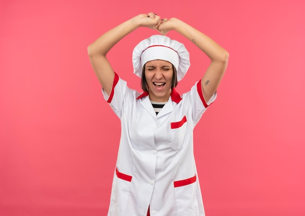 Joyeuse jeune femme cuisinière en uniforme de chef levant les poings avec les yeux fermés isolé sur rose