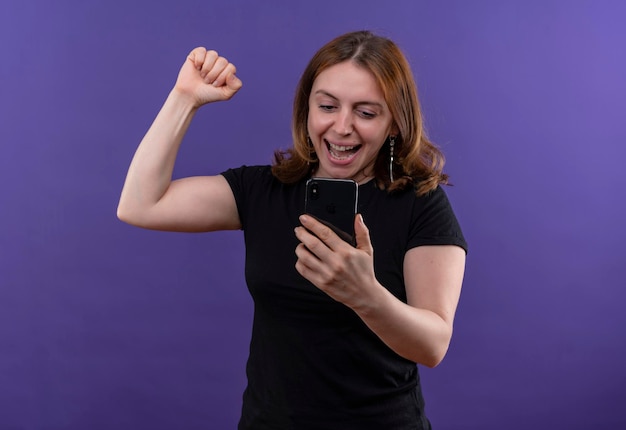 Joyeuse jeune femme décontractée tenant un téléphone mobile avec le poing levé sur l'espace violet isolé avec copie espace