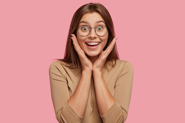 Joyeuse jeune femme européenne brune sourit positivement, garde les mains sur les joues