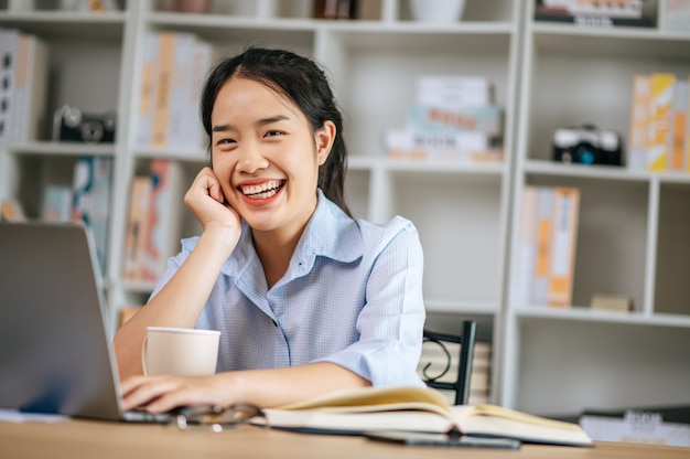 Joyeuse jeune femme jolie assise et utilisant un ordinateur portable et un manuel pour travailler ou apprendre en ligne, tenant une tasse de café à la main et souriant avec plaisir