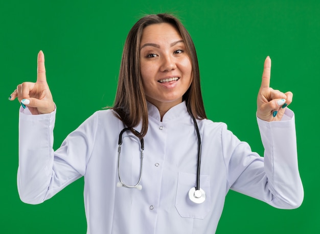 Joyeuse jeune femme médecin asiatique portant une robe médicale et un stéthoscope regardant l'avant pointant vers le haut isolé sur un mur vert