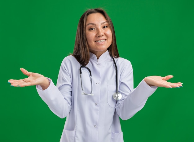 Joyeuse jeune femme médecin asiatique portant une robe médicale et un stéthoscope regardant la caméra montrant des mains vides isolées sur un mur vert