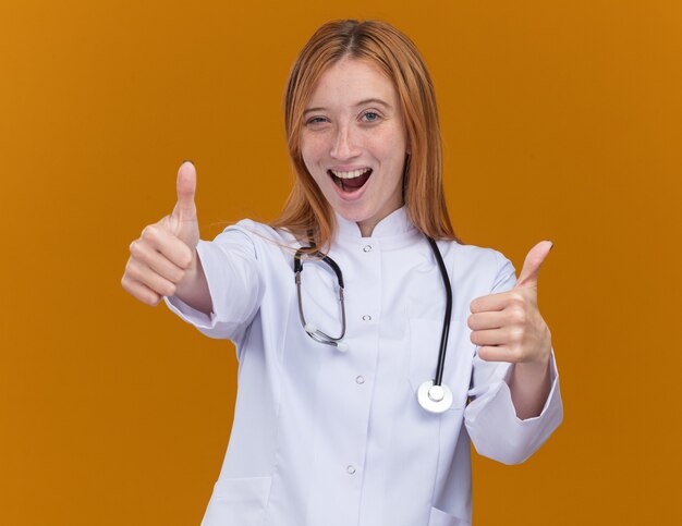 Joyeuse jeune femme médecin au gingembre portant une robe médicale et un stéthoscope regardant à l'avant montrant les pouces vers le haut isolés sur le mur orange