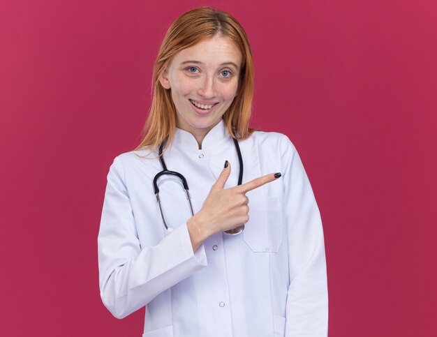 Joyeuse jeune femme médecin au gingembre portant une robe médicale et un stéthoscope regardant l'avant pointant sur le côté isolé sur un mur cramoisi avec espace pour copie