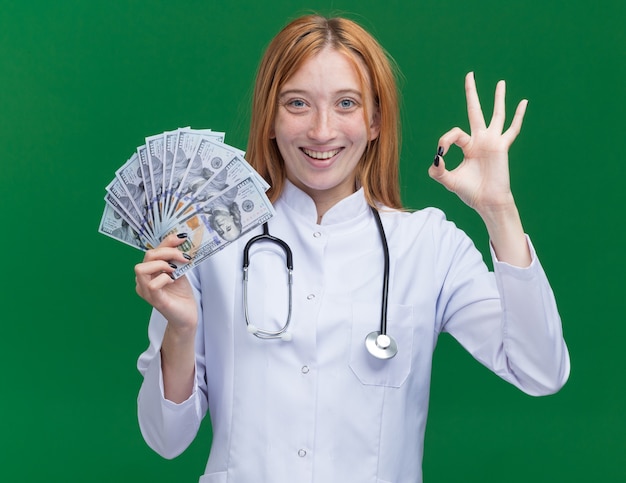 Joyeuse jeune femme médecin au gingembre portant une robe médicale et un stéthoscope tenant de l'argent faisant signe ok isolé sur mur vert