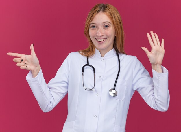 Joyeuse jeune femme médecin gingembre portant une robe médicale et un stéthoscope montrant cinq avec la main pointant sur le côté