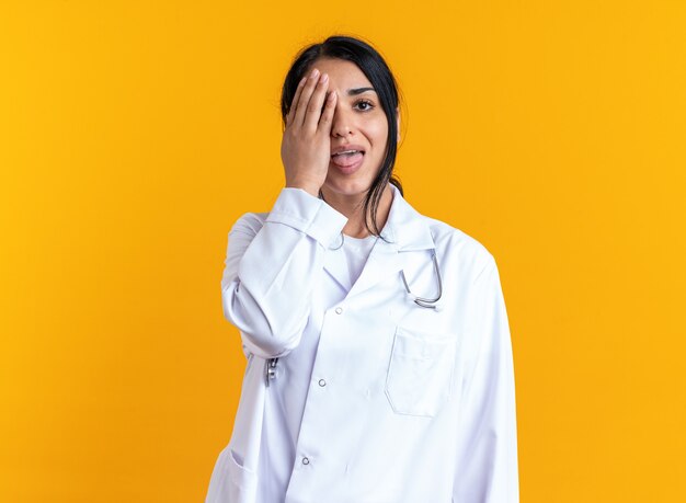 Joyeuse jeune femme médecin portant une robe médicale avec un œil couvert de stéthoscope avec une main isolée sur un mur jaune