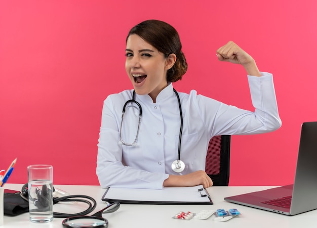 Joyeuse jeune femme médecin portant une robe médicale et un stéthoscope assis au bureau avec des outils médicaux et un ordinateur portable faisant un geste fort