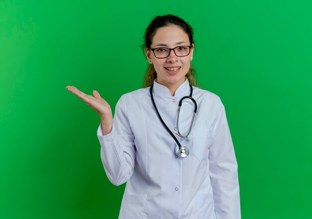 Joyeuse jeune femme médecin portant une robe médicale et un stéthoscope et des lunettes montrant une main vide isolée sur un mur vert avec espace de copie