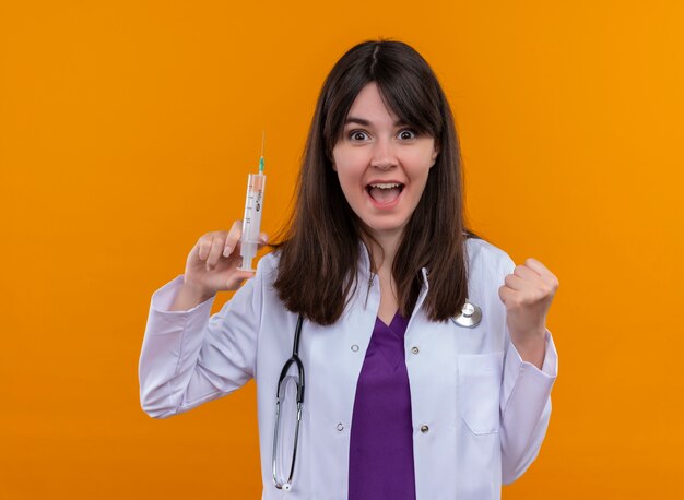 Joyeuse jeune femme médecin en robe médicale avec stéthoscope tient la seringue et garde le poing sur fond orange isolé