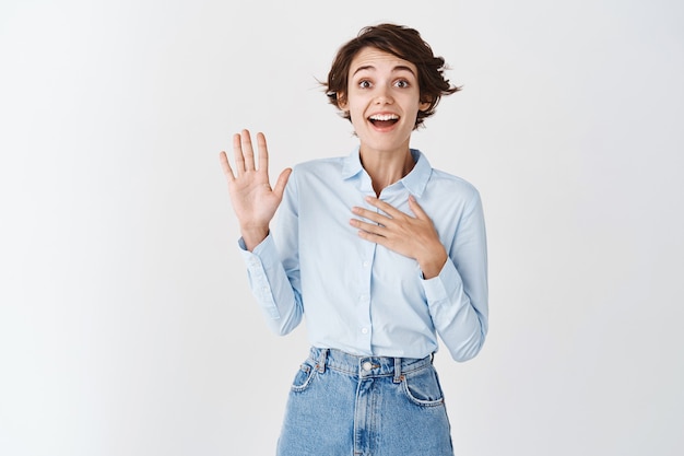 Joyeuse jeune femme naturelle levant la main, se présentant ou disposée à participer, s'auto-promotion sur un mur blanc