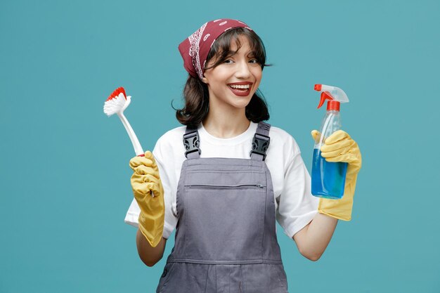 Joyeuse jeune femme nettoyante portant un bandana uniforme et des gants en caoutchouc montrant une brosse et un nettoyant regardant la caméra isolée sur fond bleu