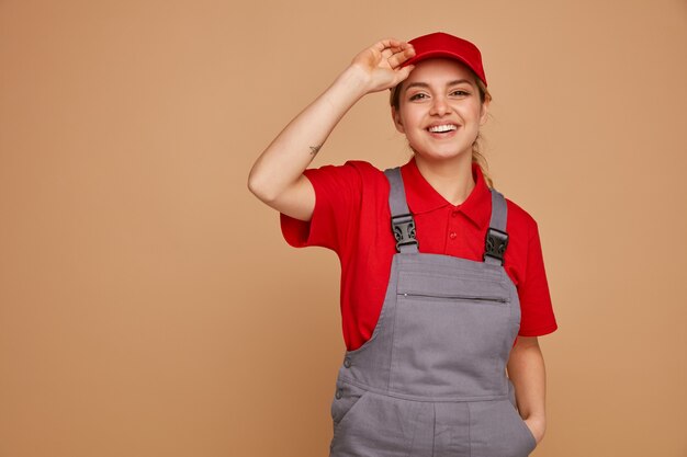 Joyeuse jeune femme ouvrier portant casquette et uniforme gardant la main dans la poche de saisie cap