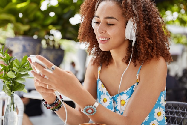 Photo gratuite joyeuse jeune femme à la peau foncée et à la coiffure afro écoute de la musique dans une liste de lecture sur un téléphone intelligent via un casque, télécharge un livre audio via une application mobile