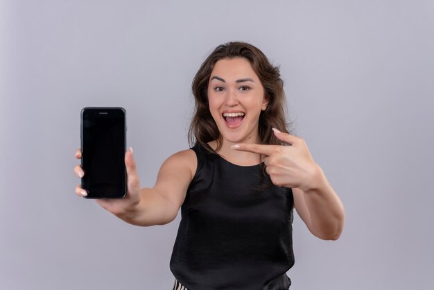 Joyeuse jeune femme portant un maillot de corps noir a tendu le téléphone pour avancer et pointer vers le téléphone sur un mur blanc