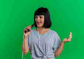 Photo gratuite joyeuse jeune femme de race blanche brune sur le casque tient le téléphone faisant semblant de chanter isolé sur fond vert avec espace de copie