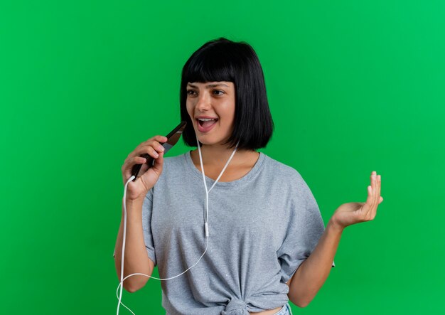 Joyeuse jeune femme de race blanche brune sur le casque tient le téléphone faisant semblant de chanter isolé sur fond vert avec espace de copie