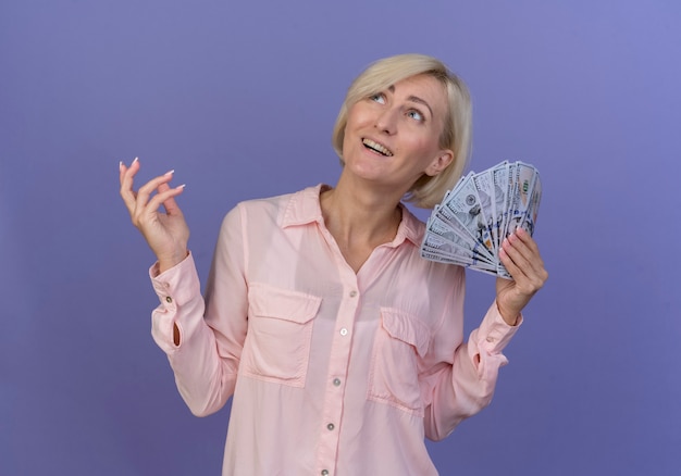 Photo gratuite joyeuse jeune femme slave blonde tenant de l'argent en gardant la main dans l'air et en levant isolé sur fond violet