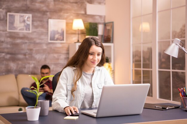 Joyeuse jeune femme souriante tout en travaillant sur un ordinateur portable depuis la maison. Petit ami se détendre sur le canapé en arrière-plan.