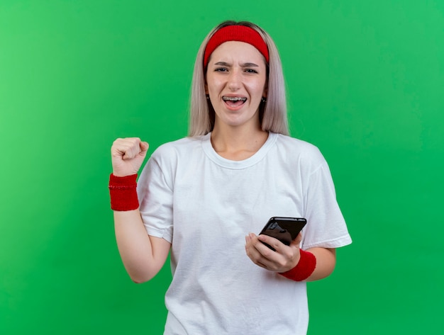 Joyeuse jeune femme sportive avec des accolades portant un bandeau et des bracelets garde le poing et détient le téléphone isolé sur le mur vert