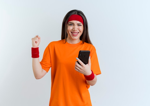 Joyeuse jeune femme sportive portant un bandeau et des bracelets tenant un téléphone mobile faisant oui geste isolé sur un mur blanc avec espace de copie