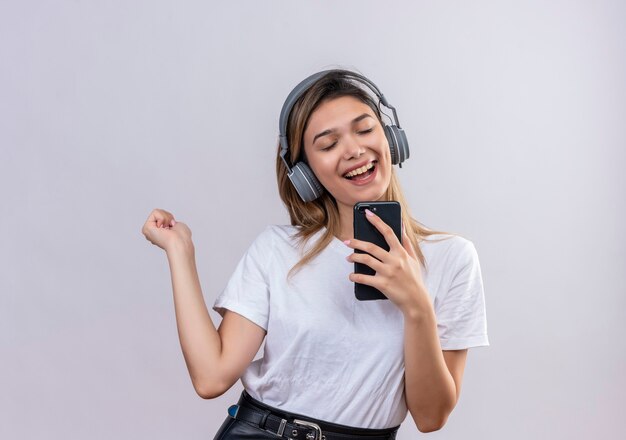 Une joyeuse jeune femme en t-shirt blanc portant des écouteurs chantant tout en écoutant la musique sur son téléphone sur un mur blanc