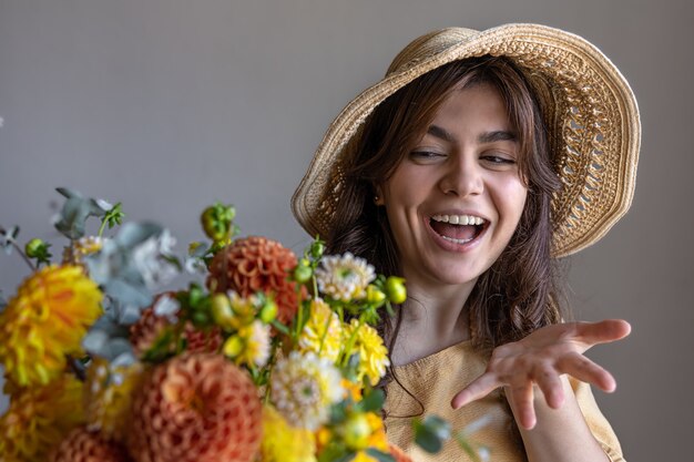 Joyeuse jeune femme vêtue d'une robe jaune et d'un chapeau avec un bouquet de chrysanthèmes lumineux et un cadeau sur fond gris.