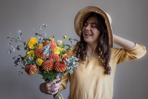 Photo gratuite joyeuse jeune femme vêtue d'une robe jaune et d'un chapeau avec un bouquet de chrysanthèmes lumineux sur fond gris.