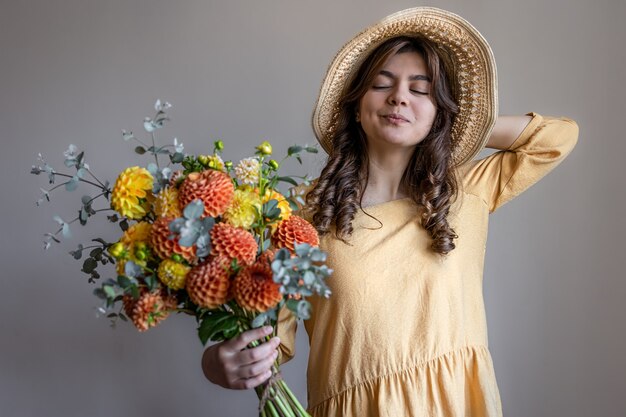 Joyeuse jeune femme vêtue d'une robe jaune et d'un chapeau avec un bouquet de chrysanthèmes lumineux sur fond gris.