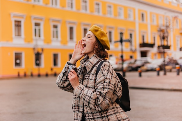 Joyeuse jeune femme voyageur appelle joyeusement quelqu'un et prend des photos sur son appareil photo rétro