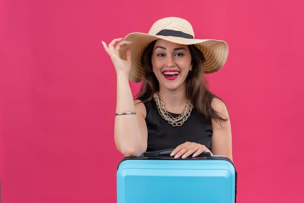 Joyeuse jeune femme voyageur portant un maillot de corps noir au chapeau a mis sa main sur le chapeau sur le mur rouge