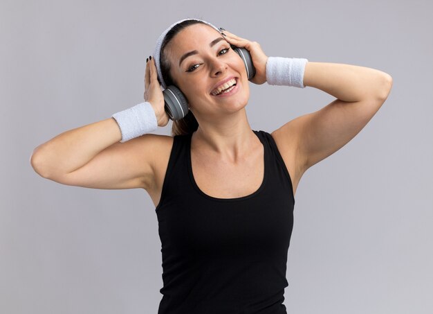 Joyeuse jeune fille assez sportive portant un bandeau et des bracelets avec des écouteurs mettant les mains sur des écouteurs écoutant de la musique isolée sur un mur blanc