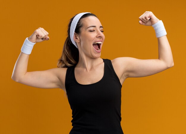 Joyeuse jeune fille assez sportive portant un bandeau et des bracelets faisant un geste fort en regardant le côté isolé sur le mur orange