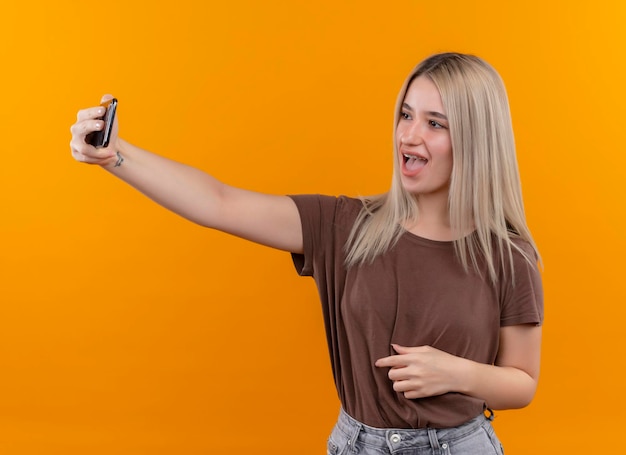 Joyeuse jeune fille blonde en appareil dentaire prenant selfie sur espace orange isolé