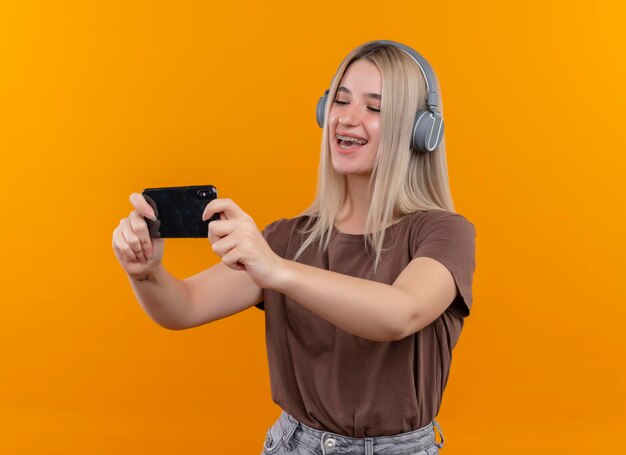 Joyeuse jeune fille blonde dans un appareil dentaire portant des écouteurs tenant un téléphone mobile en le regardant sur un espace orange isolé avec copie espace