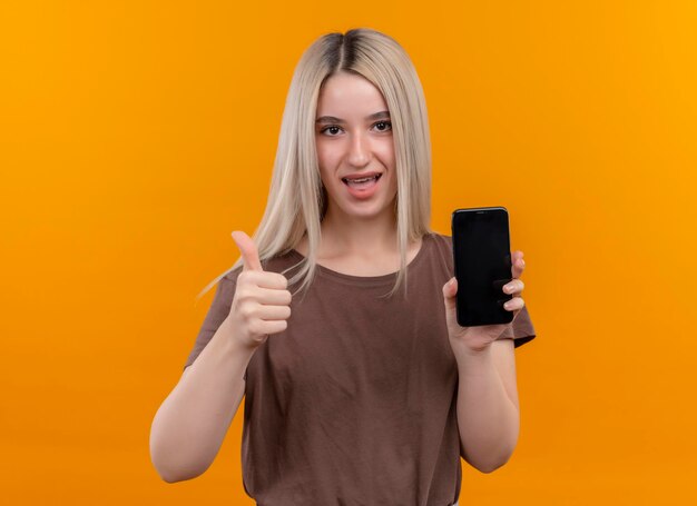 Joyeuse jeune fille blonde dans un appareil dentaire tenant un téléphone mobile montrant le pouce vers le haut sur l'espace orange isolé avec copie espace