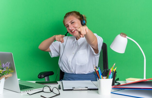 Joyeuse jeune fille blonde du centre d'appels portant un casque assis au bureau avec des outils de travail faisant un clin d'œil à votre geste