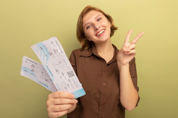 Joyeuse jeune fille blonde étirant des billets d'avion faisant signe de paix