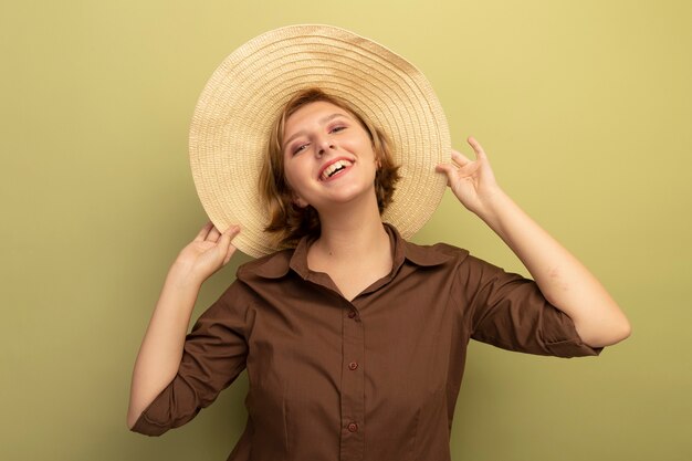 Joyeuse jeune fille blonde portant un chapeau de plage attrapant un chapeau isolé sur un mur vert olive