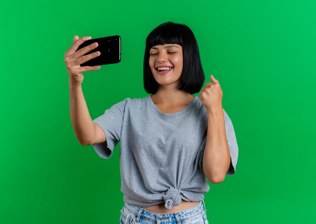 Joyeuse jeune fille caucasienne brune se tient avec les yeux fermés en gardant le poing et tenant le téléphone isolé sur fond vert avec espace de copie