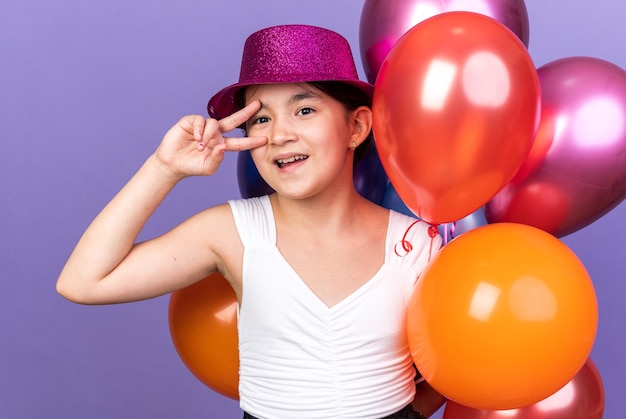 joyeuse jeune fille caucasienne avec chapeau de fête violet tenant des ballons à l'hélium et gesticulant signe de victoire isolé sur mur violet avec espace de copie