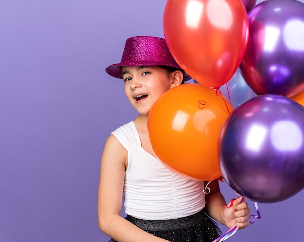 joyeuse jeune fille caucasienne avec chapeau de fête violet tenant des ballons à l'hélium isolés sur un mur violet avec espace de copie