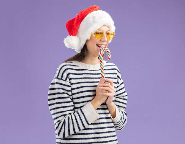 Joyeuse jeune fille caucasienne à lunettes de soleil avec bonnet de Noel tenant la canne à sucre et regardant sur le côté