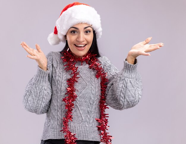 Joyeuse jeune fille caucasienne portant chapeau de Noël et guirlande de guirlandes autour du cou regardant la caméra montrant les mains vides isolé sur fond blanc