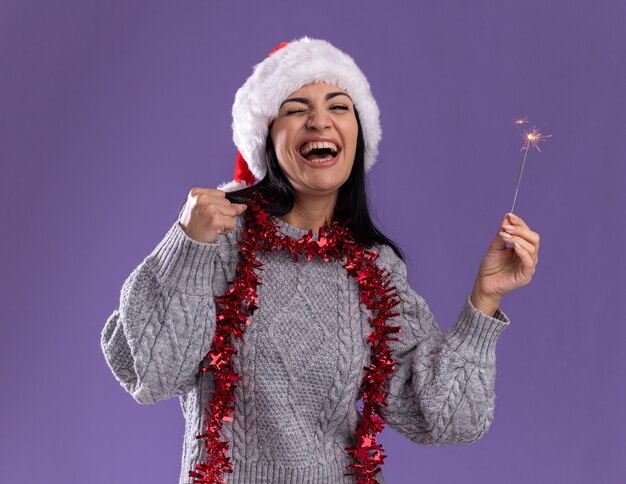 Joyeuse jeune fille caucasienne portant chapeau de Noël et guirlande de guirlandes autour du cou tenant un cierge magique faisant oui geste avec les yeux fermés isolé sur fond violet