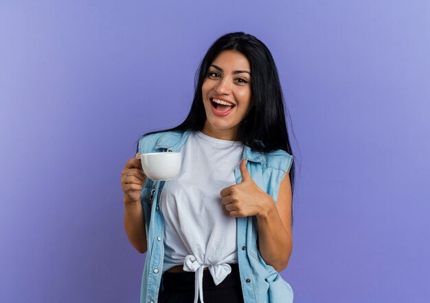 Joyeuse jeune fille caucasienne tient la tasse et les pouces vers le haut