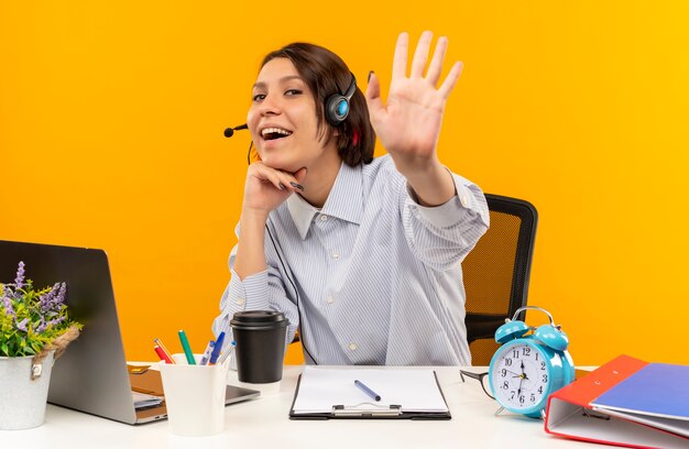 Joyeuse jeune fille de centre d'appels portant un casque assis au bureau mettant la main sous le menton et faisant des gestes salut isolé sur orange