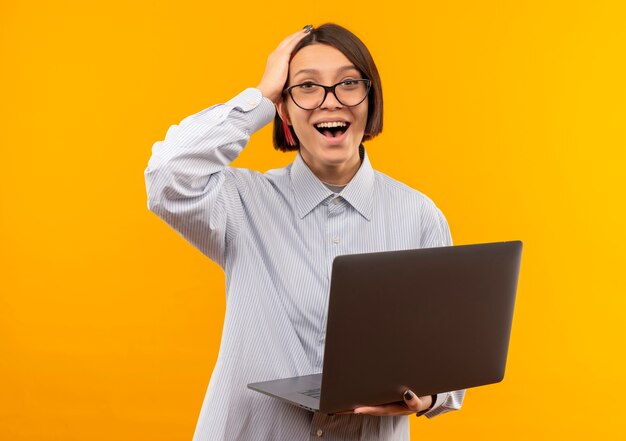 Joyeuse jeune fille de centre d'appels portant des lunettes tenant un ordinateur portable mettant la main sur la tête isolé sur orange